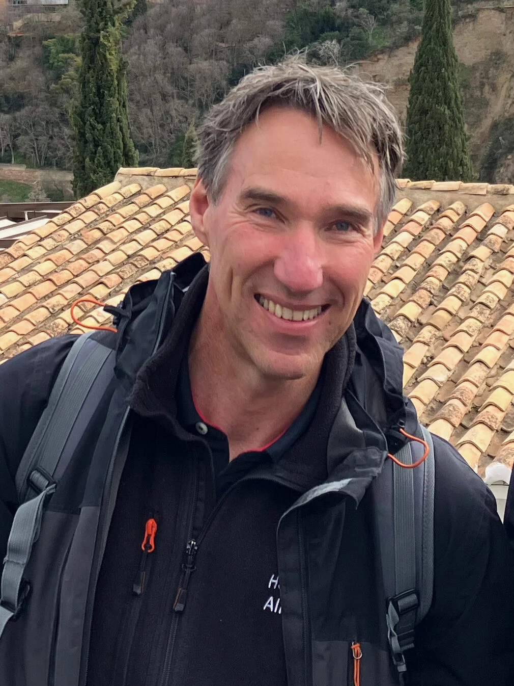 Willie met een rugzak en outdoorjas glimlacht naar de camera, met op de achtergrond een dak met terracottategels en een heuvelachtig landschap met bomen.