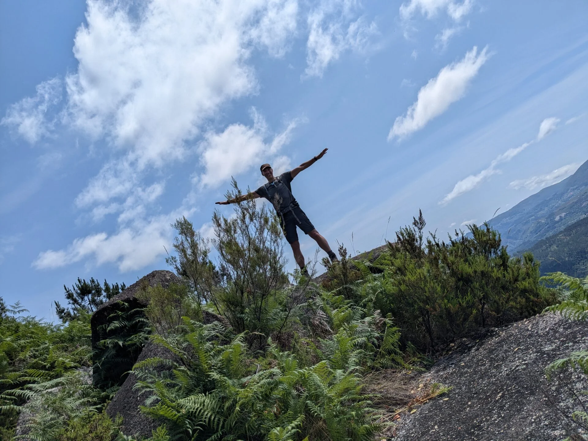 Jongeman staat met gespreide armen op een rotsachtige heuvel tussen varens en struiken, met een bergachtig landschap en blauwe lucht met wolken op de achtergrond.