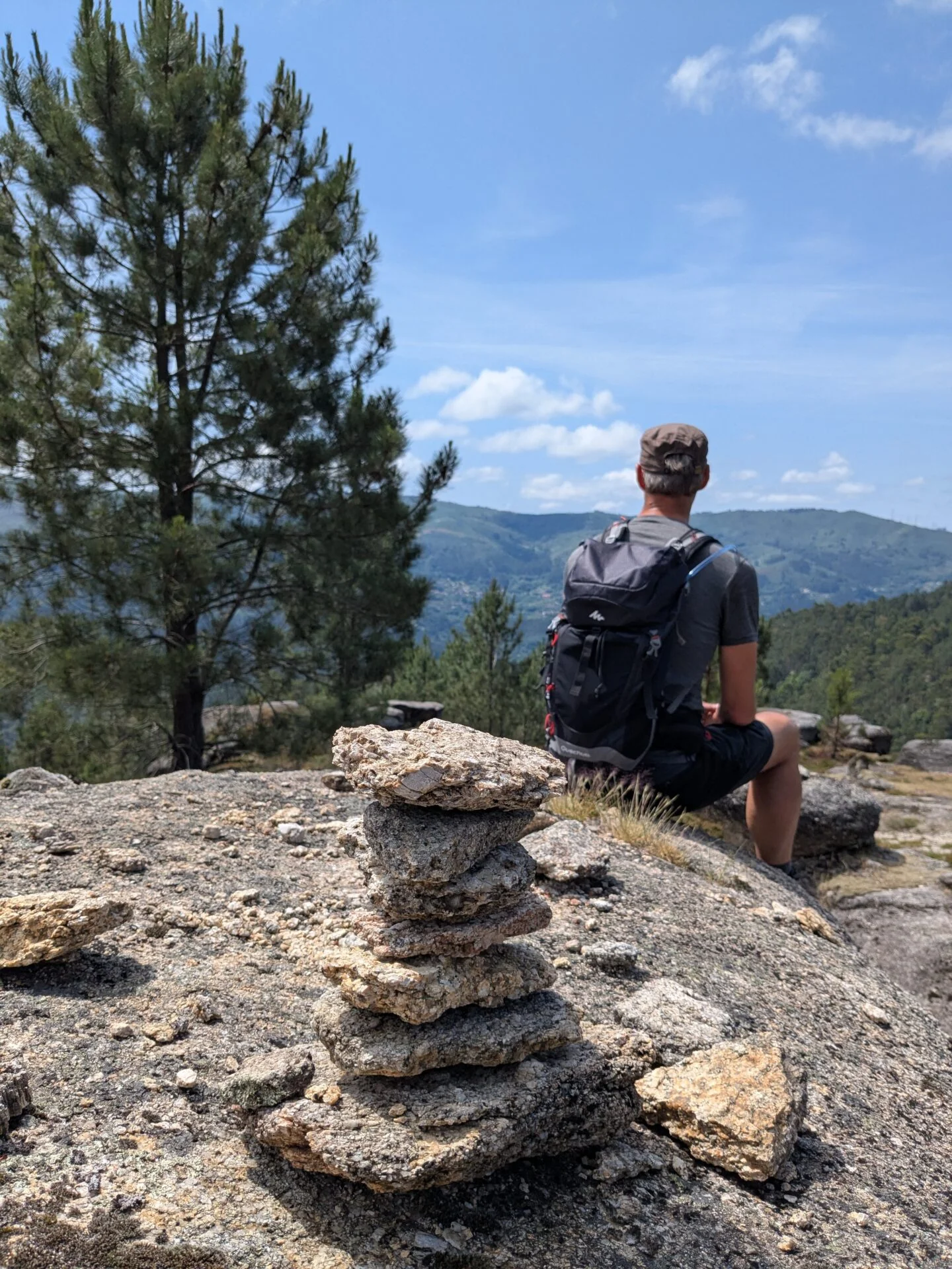 Jongeman met rugzak zit op een rots en kijkt uit over een groen berglandschap, met op de voorgrond een gestapeld stapeltje stenen en daarachter dennenbomen en heuvels onder een blauwe lucht.