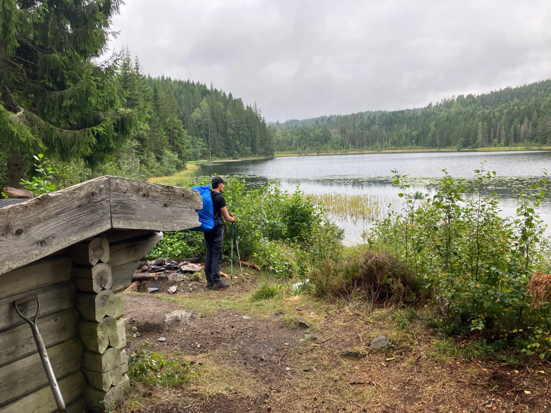 Jongeman met grote rugzak staat bij de oever van een rustig meer, naast een eenvoudige houten schuilhut, omringd door bos en water.