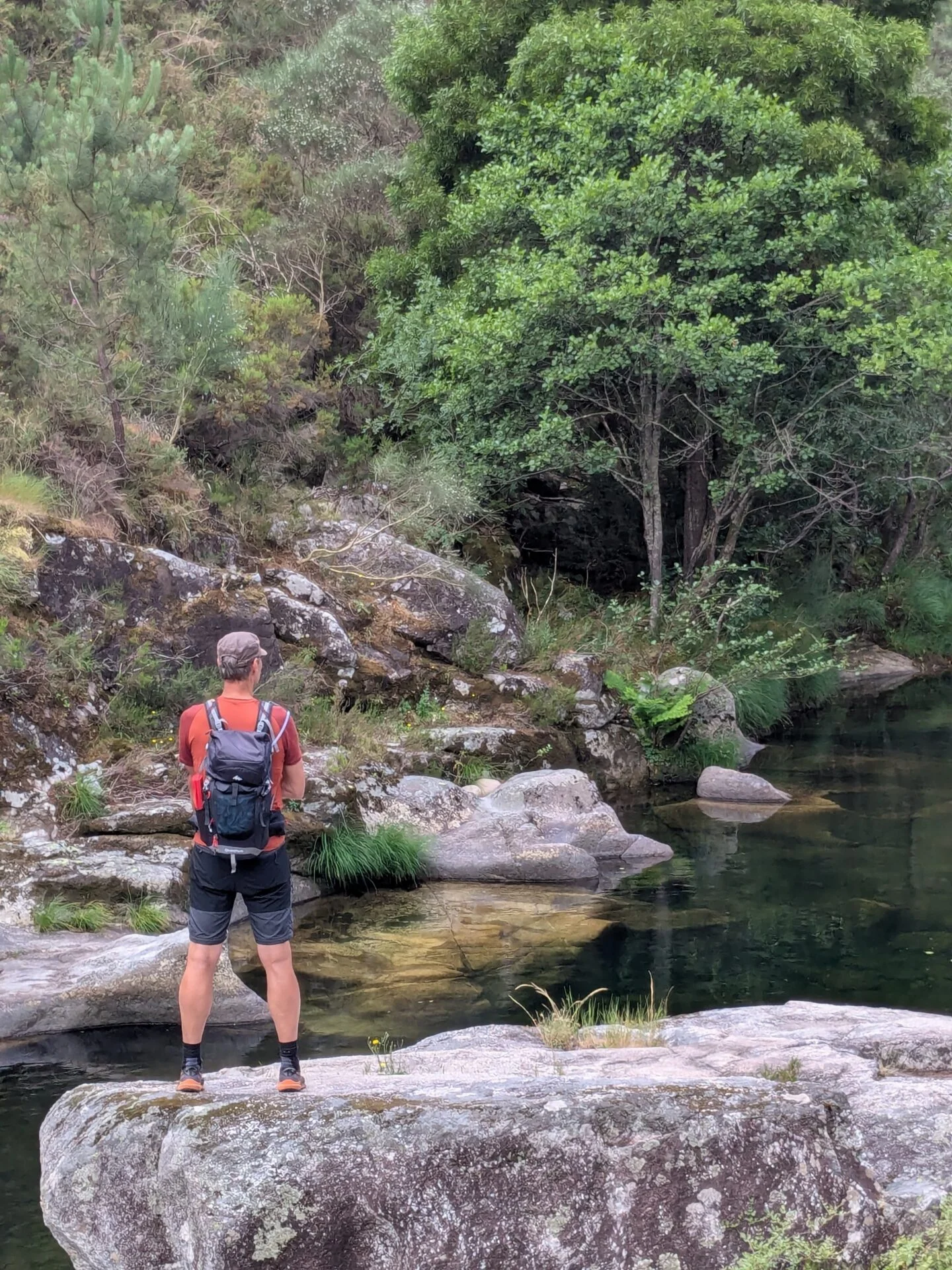 Jongeman met rugzak staat op een rots bij een heldere bosrivier, omringd door groen, stenen en bomen in een rustige, natuurlijke omgeving.