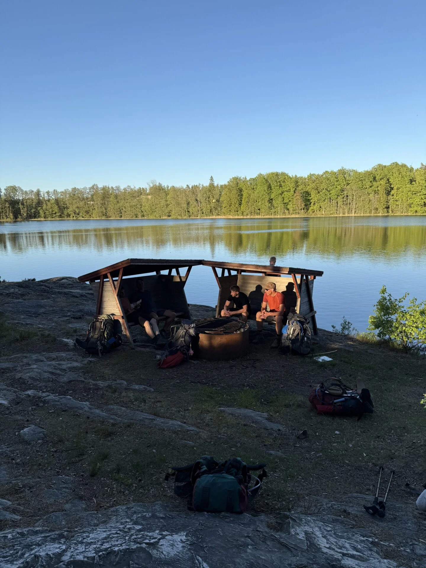 Twee mannen in gesprek rond een vuurplaats onder een houten afdakje aan de oever van een rustig meer, met rugzakken op de grond en een bos aan de overkant in warm avondlicht.